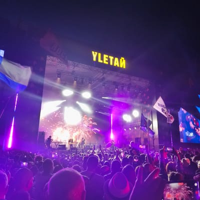 This media file depicts a lively outdoor concert scene at night, likely in Lagunovo, Russia, given the context. A large crowd of people are gathered, illuminated by vibrant purple stage lighting and the bright glow of fireworks exploding above the stage. Many in the audience are holding up their phones, capturing the spectacle.
The stage is set up with multiple lighting rigs and speakers, and a band is performing. The main focal point of the stage is a large, illuminated sign spelling out "YLETAй" in bright yellow capital letters. There are also flags visible, one white flag with text and an illustration, and a dark blue flag, both held aloft by poles. The overall atmosphere appears energetic and celebratory, with the crowd seemingly engrossed in the performance. The time of day is clearly night, suggested by the dark sky and the dominance of artificial lighting. The weather is not discernible from the image. The text "YLETAй" is prominently displayed on the stage sign. The white flag has the text "ТАМБ" on it.