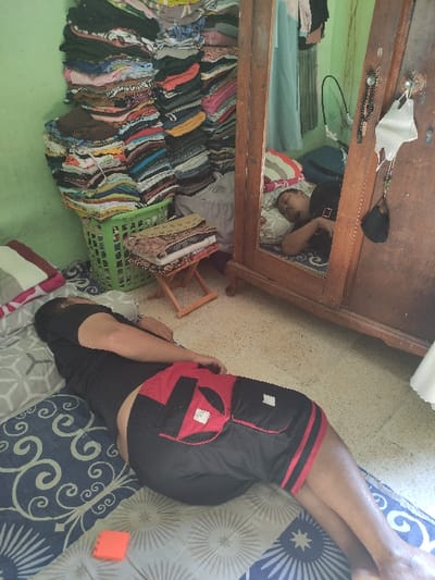 The image shows a man lying on a bed in a room in Kecamatan Lawang, Indonesia, located at latitude -7.828 and longitude 112.701. The man is wearing black shorts with a red stripe and a black shirt. His right leg is bent at the knee and resting on the bed. The bed has a blue and white patterned cover. In the background, there is an open wooden cabinet with many clothes stacked on the shelves. A green plastic basket is on the floor, with a patterned fabric behind it. There is also a small, orange object on the bed next to the man. The room has a green wall and a white tiled floor. The man's right hand is hanging off the bed, and his left hand is resting on the bed. He appears to be resting.