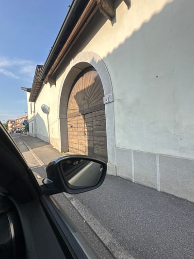 The image shows a view from the passenger side of a car driving on a street in Rudiano, Italy. The street is paved, with a sidewalk and a building on the right. The building features a large wooden arched doorway, a stucco facade, and a decorative stone base. A house number, "1", is visible on a tile next to the doorway. The sky is blue with some clouds. In the foreground, the car's side mirror reflects the passing scenery. In the distance, other buildings and some traffic are visible. The overall scene depicts a streetscape in a residential or commercial area of the town.