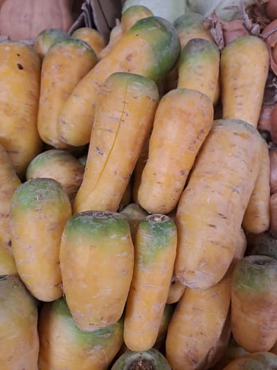 The image shows a close-up view of a pile of unwashed yellow carrots, some with green tops, likely at a market in Elektrostal, Russia. The carrots are stacked haphazardly, with varying sizes and shapes. The surface of the carrots is uneven and shows dirt, imperfections, and slight color variations. The overall lighting suggests an indoor market setting. There are no people visible.