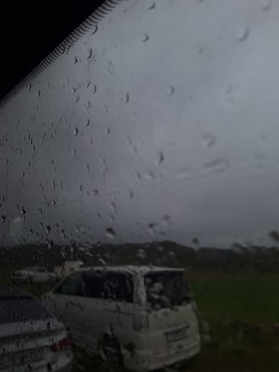 The image depicts a view through a rain-streaked windshield. The windshield wiper blade area is visible in the lower left corner. Outside, a blurred view of what appears to be vehicles parked next to a grassy area. The scene is overcast. Given the request to include the city of Tlik, Armenia, it is assumed the image was taken there, however, there is no visual information to independently confirm the location within Tlik or even that the image was taken in Tlik. No people are visible. The objects are vehicles and wet grass, the action is it is raining, the interation is one of vehicles parked in wet conditions. The environment is a gray and overcast day.