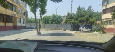 The image depicts a street scene in Andijan City, Uzbekistan, on a clear day. In the foreground, a car's dashboard is visible with a booklet resting on it. The main scene shows a light-colored, four-door hatchback parked on the right side of the street. Across the street, a multi-story residential building with yellow and red sections dominates the left side of the frame, featuring numerous windows and air conditioning units. A dark-colored station wagon is parked in front of this building. Further down the street, a gray brick wall runs along the property line, with yellow pipes visible on top. A street lamp with a curved pole stands near the wall. Several trees, including deciduous and coniferous varieties, are scattered throughout the scene, some casting shadows on the asphalt road and concrete ground. A tall communication tower is visible in the distance against the bright sky. The environment appears to be an urban residential area, with a mix of buildings, vehicles, and vegetation.