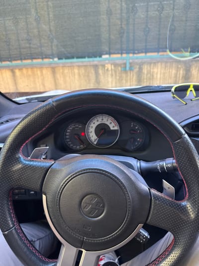 The image shows the interior of a car, focusing on the steering wheel and dashboard. The steering wheel is black with red stitching and has paddle shifters. The dashboard includes a speedometer and tachometer. A pair of neon yellow sunglasses is resting on the dashboard. Outside the car, a black mesh fence is visible above a concrete wall. The scene suggests the car is parked outdoors in a location with a fence and wall. The sunglasses indicate a sunny environment. There are no people clearly visible in the image, but the perspective suggests the photograph was taken from the driver's seat. The overall impression is of a parked car, possibly on a warm day.