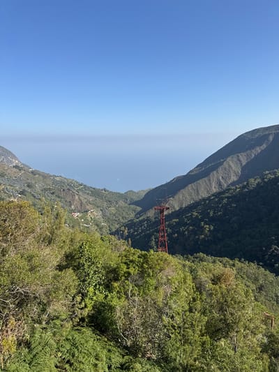 The image shows a view in the city of San Antonio de Galipán, Venezuela. In the background, there is a tall mountain and a deep ravine with a red metal structure (likely a part of a tram or cable car system) spanning across it. The foreground is filled with dense green vegetation, including trees and shrubs. The sky is a clear blue with a slight haze near the horizon. The photo is taken during daylight, evidenced by the lighting. There are no people visible in the image.