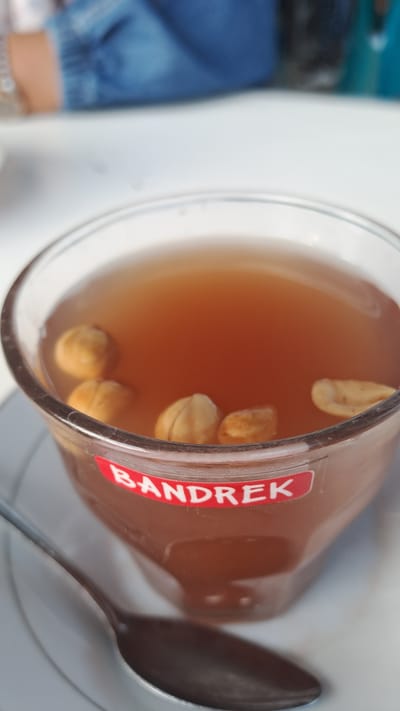 A close-up, eye-level shot shows a clear glass mug filled with a dark amber liquid, possibly a beverage like bandrek, as indicated by a red and white label on the glass that reads "BANDREK." Four light brown, round objects, resembling nuts or seeds, are visible floating in the liquid. The mug rests on a white saucer, and a dark metal spoon is positioned to the left of the mug, partially submerged in the saucer. The background is blurred, but a person wearing a blue denim jacket is partially visible, suggesting a casual setting. The scene appears to be indoors, possibly in Tingkeummanyang, Indonesia, where such beverages are commonly consumed.