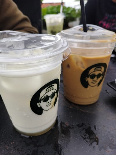 This close-up shot captures two iced beverages in clear plastic cups placed on a dark, possibly metallic or stone, table in an outdoor setting in Dramaga, Indonesia. The cup on the left contains a light-colored, possibly milky, drink with ice, while the cup on the right holds an iced coffee with a black straw protruding from its clear dome lid. Both main cups feature an identical black and white logo: a cartoonish illustration of a person's head wearing a backwards baseball cap and sunglasses. The table surface beneath the cups shows signs of condensation or moisture, hinting at cold drinks or a humid environment. In the softly blurred background, a person's brown-sleeved arm is visible alongside lush green foliage and another distant plastic cup with a green-tinged beverage, suggesting a cafe or casual eatery. The natural, bright lighting indicates it is daytime. No discernible text is present, apart from the distinctive visual logo on the cups.