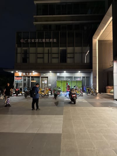 The image depicts a nighttime scene outside a modern building in Huangpu District, China. The building features a dark glass facade with the text "广东省湖北襄阳商会" (Guangdong Hubei Xiangyang Chamber of Commerce) visible in pink neon. Below this, a storefront with an orange awning displays the text "壹号咨询" (No. 1 Consultation).
In front of the building, a paved area is occupied by several electric scooters and bicycles, some of which are parked haphazardly. Two men are present. One, wearing a black t-shirt and shorts, is walking with a white bag in his hand. The other man, in a blue t-shirt and dark pants, is looking at his phone, with a backpack on. To the right, a person in a red helmet and vest is interacting with a group of parked bicycles. Several boxes and a green bag are stacked near the building's entrance on the far right. The overall atmosphere suggests a commercial or service area that operates into the evening.