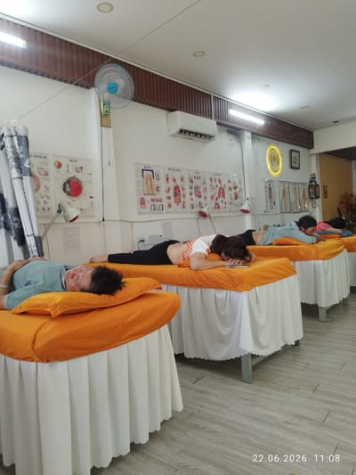In a room with white walls and a wooden ceiling, four people are lying face down on massage beds covered with orange sheets. The room appears to be a wellness center or clinic. Medical charts depicting anatomical diagrams of the human body, including the eye, teeth, and circulatory system, are displayed on the wall. A wall-mounted fan is visible near the ceiling, and an air conditioner is mounted above the charts. There are also several lamps with red bulbs positioned near the beds, possibly for heat therapy. The setting suggests a tranquil environment for relaxation or treatment. The date stamp at the bottom right corner of the image reads "22.06.2026 11:08", indicating the time of day. The overall mood is calm and still, with individuals seemingly resting or receiving a treatment. The location context is Binh Thuan Ward, Vietnam.