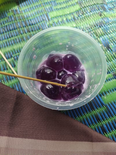 A clear plastic cup filled with dark purple, spherical objects in a light purple liquid sits on a textured brown cloth. A thin wooden skewer is inserted into one of the purple objects. The cup is placed on a woven mat with alternating blue and green stripes. The scene is indoors, likely on a table or flat surface, with no indication of the time of day or weather. The focus is on the food item in the cup, suggesting it might be a dessert or snack. There is no visible text in the image.