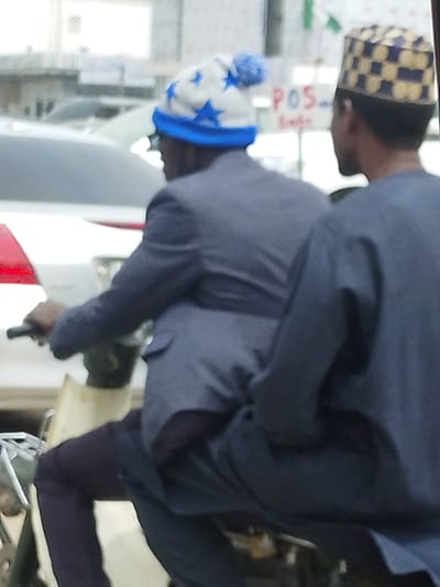 The image depicts two males riding a motorcycle or scooter on a street in Gyadi Gyadi Arewa, Nigeria, during daylight. The individual in the driver's position, viewed from behind on the left, wears a grey suit jacket, dark trousers, and a white knitted beanie adorned with blue stars, a blue brim, and a blue pom-pom. His left hand is visible on the vehicle's handlebar. A passenger is seated directly behind the driver, seen from the back on the right. This individual wears a dark blue long-sleeved garment and a yellow and blue checkered cap. The vehicle appears to be light-colored, possibly off-white. In the blurred background, other white vehicles are discernible, and a white sign with "POS" in red lettering is visible to the right. Faint green and white shapes, possibly flags, are also present in the distance.