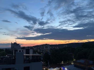 The image depicts a dusk cityscape, likely in Pernik, Bulgaria, with a vibrant sunset illuminating the sky. The foreground shows rooftops of buildings, some with solar panels and decorative blue lights. Mid-ground features apartment blocks, trees, and a prominent tall building topped with antennas. The background is dominated by a silhouette of hills and a few scattered lights from houses. The sky is a gradient of blues and grays, transitioning to warm orange and yellow hues near the horizon, with scattered clouds. No people are visible in the frame.