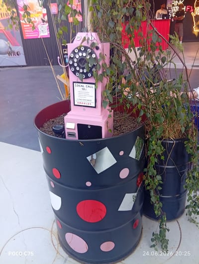 This media file features a close-up shot of a pink rotary dial telephone, repurposed as a decorative element, placed within a large black planter. The telephone is adorned with a sign that reads "LOCAL CALL 10¢" and provides instructions for its use, including "Remove Handset," "Wait for dial-tone," "Wait for party to answer," and "Drop in coin," along with information for "LONG DISTANCE." The planter itself is decorated with scattered circular and square reflective elements. Lush green foliage from a plant cascades over and around the telephone and planter, creating a natural, somewhat overgrown aesthetic.
In the background, hints of an outdoor venue are visible. A bar with a red counter is present, with two individuals standing behind it. To the left, a poster with the text "WRG" is partially visible. Neon lights and other signage suggest a vibrant, possibly entertainment-focused location. The overall scene appears to be outdoors, given the natural light and the presence of plants.
The timestamp at the bottom right corner indicates the image was taken on "24.06.2026 20:25," suggesting an evening setting. The visual elements, particularly the bar and signage in the background, imply a social gathering or a public space like a beer garden or an outdoor festival. No specific emotions are conveyed by the subjects, as the focus is primarily on the decorative telephone and planter. The location context of Wrocław, Poland, is provided separately.