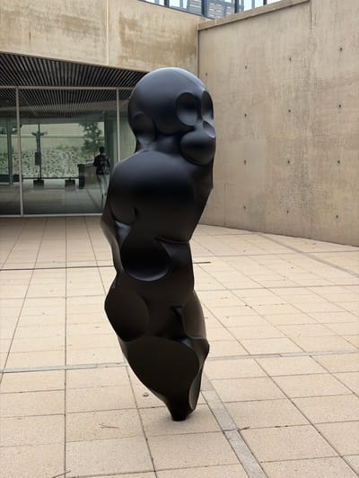 A large, black, abstract sculpture with an organic, bulbous form and several circular indentations is positioned outdoors on a light-colored tiled surface. The sculpture is the primary subject of the image. In the background, through a large glass window or storefront, reflections show a person and trees. The building appears to be modern, with concrete walls and horizontal slatted elements. The lighting suggests it is daytime, with shadows cast on the tiled ground. The overall setting is an urban or architectural environment.