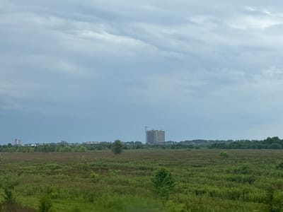 The image depicts a broad, flat landscape dominated by a vast expanse of tall green grass and brush. In the distance, a low-lying treeline stretches across the horizon, interrupted by several buildings and what appears to be a construction site with a tall, unfinished structure. The sky above is overcast with a gradient of blue and grey clouds. The scene suggests a rural or semi-developed area on the outskirts of a city.