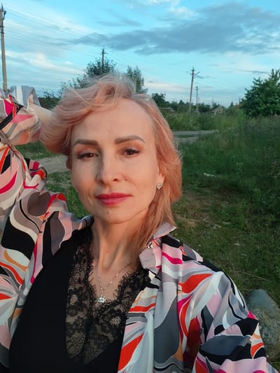 A woman with light pink hair, wearing a black lace-trimmed top and a colorful patterned shirt, poses for a selfie outdoors. She has on makeup and a silver necklace with a crown pendant, and a heart-shaped earring on her left ear. The background shows a grassy area with trees and utility poles, suggesting a rural or suburban setting. The sky is blue with some clouds. The image appears to be taken in Vasil'ki, Russia.