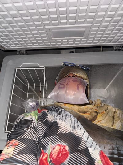 The image depicts a person lying inside an open freezer, their face covered by a plastic bag filled with liquid. The person is wearing glasses perched on their head and is wrapped in patterned fabrics. The freezer is white with a wire basket inside. In the background, there are bags of what appear to be ice or water. The scene is domestic, suggesting a personal and unconventional cooling method. The location of Yelwa, Nigeria, is provided, but no visual cues in the image confirm this specific location.
