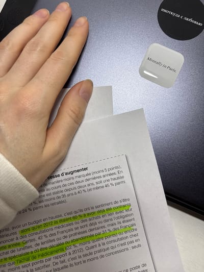 The image presents an overhead close-up view of a human left hand partially resting on a gray electronic device, likely a laptop or tablet, in Kudrovo, Russia. A white sheet of paper with printed French text is overlaid on the device, occupying the lower-left and central portions of the frame. Sections of the French text, discussing percentages related to medical consultations and medication purchases, are highlighted in yellow. The hand displays light skin and natural, unpainted fingernails, with a white sleeve visible on the wrist. Two stickers are affixed to the upper-right surface of the electronic device: a black circular sticker with white Cyrillic text reading "ниоткуда с любовью" (from nowhere with love), and a glossy white rectangular sticker with rounded corners displaying black English text, "Mentally in Paris."