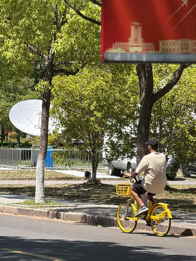 A man on a yellow bicycle is riding on a paved road in the Jiangning District, China. The man is wearing a light-colored shirt and a baseball cap. The bicycle has a basket in the front. A tree is visible to the left of the man, and a satellite dish is mounted on a tree trunk nearby. A red sign is partially visible on the left edge of the frame. A sidewalk and a grassy area with trees are to the left of the road. Cars are parked on the street nearby.