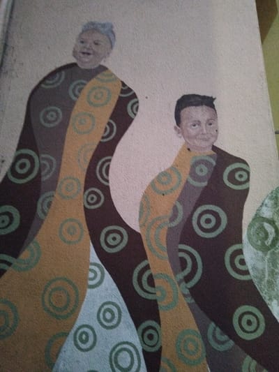 This media file shows a mural on a wall, likely a public space given the subject matter and style. The mural depicts two figures, possibly children or stylized representations of people, lying down. They are rendered in a somewhat abstract style, with elongated bodies and simple facial features. The figures are covered in patterned fabrics or flowing shapes, featuring concentric circles in shades of green, brown, and yellow. The background is a textured, light-colored wall. One figure appears to be older, with darker hair and a more defined face, while the other is younger with a lighter complexion and a more simplistic depiction of features. The overall impression is one of peaceful repose or a dreamlike state. There are no visible signs of a specific time of day, weather, or immediate location cues beyond the mural itself. The art style suggests a modern, possibly folk-inspired aesthetic. The context provided indicates this mural is located in Vila Nova de Famalicão, Portugal.
