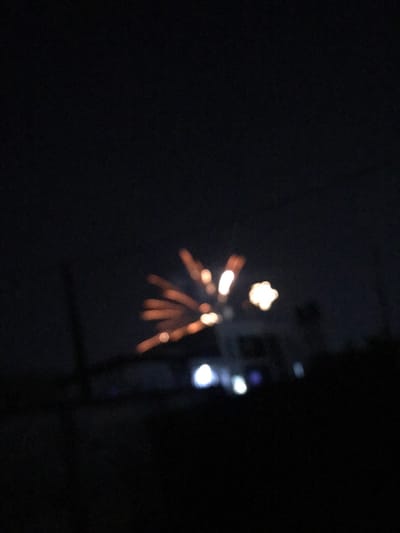 In a dark night scene, a blurry burst of light resembling fireworks appears. The light radiates outwards, featuring a central, star-shaped glow with smaller, linear trails extending downwards and outwards. The image is out of focus, lending to a hazy quality. Adjacent to the burst are additional blurred points of light, possibly streetlights or other distant sources. The surrounding area is enveloped in darkness.