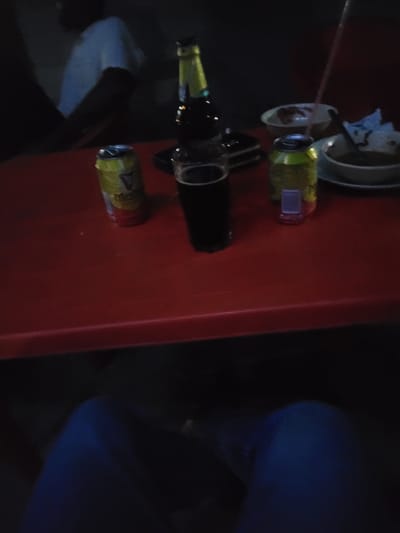 The image depicts a dimly lit scene at a location in Keffi, Nigeria. A red rectangular table occupies the foreground and midground. Underneath the table, a person's legs, clad in blue trousers, are visible. In the background, to the upper left, a person is partially visible from the chest upwards, wearing a light-colored top, with their face obscured by darkness.
On the red table, multiple items are arranged:
- To the left, an open yellow aluminum can, bearing a partial "GUINNESS" logo, is present.
- Adjacent to it, a clear glass filled with a dark liquid (consistent with stout or dark beer) is visible.
- Behind the glass and can, a brown glass bottle with a yellow and green label and cap is positioned.
- Further to the right, a second yellow aluminum can, similar to the first, is also open.
- Between these two cans, a small, blurry, purple object, possibly a miniature bottle or container, rests on the table.
- On the right side of the table, two white bowls are present. The rear bowl contains a light-colored substance, and a pink drinking straw is inserted into it or nearby. The front bowl contains a darker, viscous substance, possibly food, with a crumpled light-colored napkin partially submerged or resting on its edge.
The arrangement of beverages and food suggests a casual social gathering or meal in a low-light environment, likely an evening or nighttime setting.