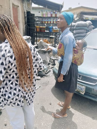 The image portrays an outdoor street scene in Kadoma, Zimbabwe, under bright daylight. In the foreground, facing away from the camera, is an individual with long, light brown braided hair, wearing a white shirt with a black heart pattern and light-colored trousers. To their right, a woman stands facing left, wearing a light blue long-sleeved shirt, a dark skirt, and light-colored flip-flops. Her head is covered by a turquoise headwrap. A baby, appearing to be asleep, is secured to her back with a colorful, patterned fabric wrap. The woman's right arm is slightly raised, with her palm open, suggesting a gesture while speaking, indicated by her partially open mouth. She holds a small, light-colored object in her left hand, which rests by her side.

Behind the woman and child, a light green sedan is partially visible, facing the viewer, with a license plate displaying characters '09AE'. Further into the background, an outdoor market stall is present, featuring a metal rack holding multiple plastic bottles containing various colored liquids, along with several plastic buckets. Stacked grey sacks are piled behind the car. The ground is paved with asphalt, exhibiting some cracks and scattered small debris. Rough-textured, light-colored buildings with closed doors form the backdrop of the scene. Strong sunlight illuminates the area, casting distinct shadows on the ground.