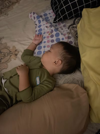 A young baby or toddler is captured sleeping peacefully on a bed in an indoor setting in Ngasinan, Indonesia. The child, lying on their side with closed eyes, wears a long-sleeved green sleeper or onesie with buttons down the front. A small blue tag on the sleeper's chest clearly displays the text "baby cute." A thin, dark bracelet adorns the baby's right wrist.

The baby's head rests on a small white pillow decorated with blue and purple polka dots, featuring a Snoopy character design with visible "SNOOPY" text. Surrounding the sleeping child are several other pillows: a large, light yellow one at the top and a large, light brown/beige one to the right, possibly a body pillow. The bed is covered with a light-colored sheet that has an intricate patterned border. A dark fabric with a white grid pattern is visible on the far left, suggesting another pillow or blanket.

The dim lighting suggests it is either nighttime or naptime. The scene conveys a sense of calm and tranquility, with the baby appearing deeply and peacefully asleep.