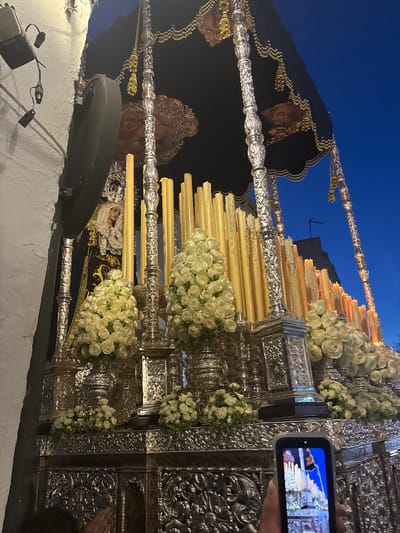 The image shows a religious procession, likely a Holy Week event, in Córdoba, Spain. A richly decorated platform, adorned with silver embellishments, candles, and bouquets of white roses, is the focal point. On the platform, religious figures, likely depictions of the Virgin Mary and Jesus Christ, are visible. The figures are draped in ornate clothing. One person is holding a mobile phone, recording the scene. The background consists of a dark blue sky, suggesting the event is taking place at night. A building is visible between candles. A street light is visible below the platform.