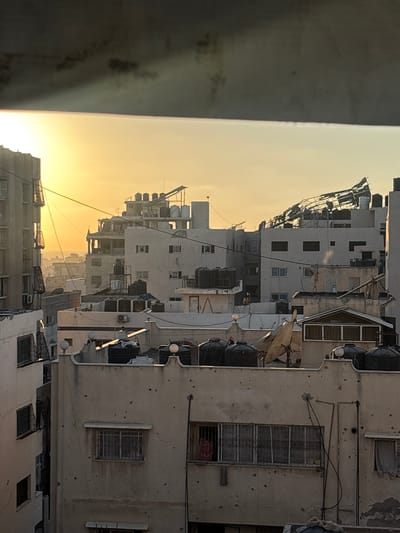 This photograph, taken from a high vantage point, offers a view of the urban landscape of Gaza City, Palestinian Territories. The scene is illuminated by the soft, warm light of a sunset, casting long shadows across the rooftops. Dominating the view are numerous multi-story buildings, many of which exhibit signs of damage. Several structures appear to have sustained significant structural compromise, with exposed beams and debris visible on rooftops. Numerous black water tanks are strategically placed on the roofs of most buildings, suggesting a reliance on stored water. The walls of the buildings are pockmarked with visible holes, indicative of shelling or impacts. The windows of many buildings are barred or covered with screens, and some windows are damaged or boarded up. In the foreground, a large satellite dish is visible on a rooftop, alongside more water tanks. The overall impression is one of a densely populated urban environment that has endured conflict. There are no individuals clearly visible in the photograph. The sky is a gradient of pale yellow and orange, transitioning to a dusky gray at the top of the frame, where a dark, ominous cloud formation is present.