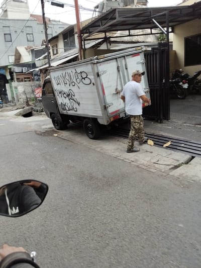 A man wearing a white t-shirt, camouflage pants, and a baseball cap is walking near a white truck with graffiti on its side. The truck is parked on a street, and there are motorcycles parked in the background. The scene appears to be in an urban area, possibly in Bandung, Indonesia, given the provided location context. The ground is paved, and there are some drains or grates on the side of the road. The lighting suggests it might be daytime, but the sky is overcast. The man seems to be engaged in some activity related to the truck, but it's unclear what he's doing. The graffiti on the truck is illegible. The overall atmosphere is casual and urban.