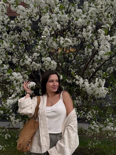 A young woman poses with a blossoming tree. She wears a white tank top, a light-colored textured jacket, and a crossbody bag. She holds a cluster of white blossoms in her hand. The tree is in full bloom with small white flowers. In the background, a dark, possibly mirrored, building facade is visible. The scene appears to be located in Odintsovo, Russia, suggested by the context of the surrounding area.