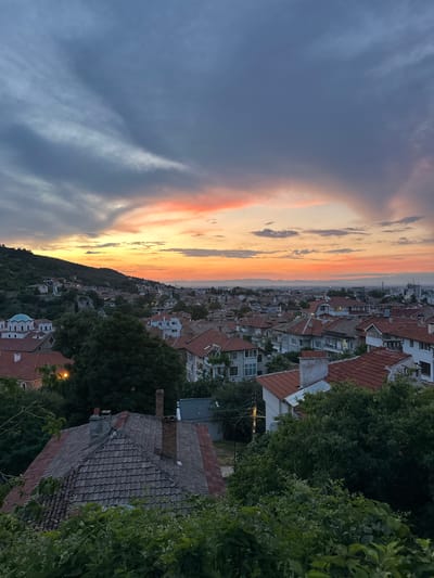 This is a high-angle, medium shot of Asenovgrad, Bulgaria, taken during sunset. The sky is filled with dramatic clouds, transitioning from dark grey to vibrant oranges and pinks near the horizon. Below the sky, the cityscape is visible, characterized by a dense cluster of buildings with red-tiled roofs. Many of these buildings appear to be residential houses.
To the left of the frame, a green, tree-covered hillside rises above the city. On this hillside, several structures are visible, including a building with a distinctive blue dome and white walls, likely a church or a similar religious building. The foreground is dominated by lush green foliage and the roofs of houses, creating a layered effect and framing the view of the city. The lighting suggests it is late afternoon or early evening, with a warm, soft glow illuminating the scene. The overall impression is one of a tranquil evening in a historic Bulgarian town.