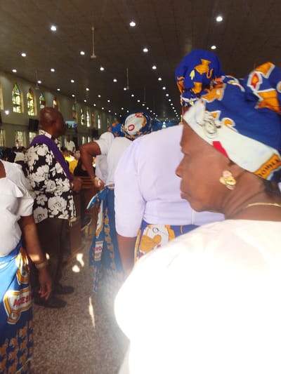 The image displays an interior scene within a large building, identified as being in the city of Sabon Gari West, Nigeria. Multiple dark-skinned individuals are standing within what appears to be an aisle or open area.

The building features a light-colored, flat ceiling equipped with numerous evenly spaced recessed circular lights and several white ceiling fans. The upper sections of the walls contain multiple arched stained-glass windows displaying patterns predominantly in yellow, green, and red hues. The floor is tiled with a light-colored surface. Dark wooden benches or pews are partially visible in the mid-ground and background.

In the immediate foreground on the right, the right profile of a dark-skinned individual is visible. This person wears a blue, white, and orange patterned headwrap, a light-colored top, a gold-colored earring, and a gold-colored necklace. Behind this individual, another person is seen from the back, wearing a light purple top and a blue, white, and yellow patterned skirt or wrapper, topped with a blue and orange headwrap. To the mid-left, a dark-skinned male individual is present, wearing a black and white floral short-sleeve shirt, dark trousers, and a purple stole draped over his shoulders; he is oriented towards the left. To the far left, a partial view of another dark-skinned individual is seen, wearing a white top and a blue patterned skirt or wrapper. Other indistinct individuals are visible further back within the structure. The scene is brightly illuminated by both natural light from the windows and artificial light from the ceiling fixtures.