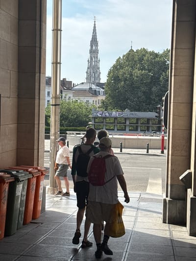 The image depicts a group of four individuals walking outdoors on a sunny day. They are walking away from the camera, with their backs mostly to the viewer, in a European urban setting. The individuals appear to be casually dressed, with two of them carrying backpacks. One person in the foreground is wearing a white t-shirt and shorts, and another is wearing a similar outfit with a baseball cap. The building architecture suggests a European city, and the tall spire in the background is a strong indicator of Brussels, Belgium, possibly the Brussels Town Hall. The time of day appears to be daytime, with bright sunlight casting shadows, indicating clear weather. The scene takes place on a paved sidewalk next to a street with traffic. In the lower right corner of the image, several large cylindrical objects, likely trash or recycling bins, are stacked. No visible text is clearly legible on the people or objects, except for some graffiti on a structure in the background. The overall mood appears casual and relaxed, consistent with people sightseeing or going about their day in a city.