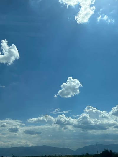 The image captures a wide-angle view of a sky filled with white, fluffy clouds against a bright blue backdrop. In the foreground, a range of dark, silhouetted mountains stretches across the horizon. Below the mountains, a strip of land is visible, with sparse vegetation and a single lamppost on the right side. The overall scene suggests an outdoor environment in a rural or semi-rural area, possibly in or around Shtarkovo, Bulgaria, during daylight hours. There are no people or specific structures that would indicate an urban setting.