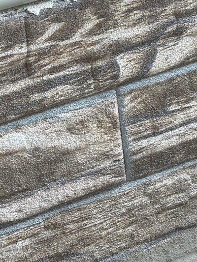 The image shows a close-up view of a textured surface, resembling faux stone or brickwork with a rustic, weathered appearance. The material is a granular, multi-toned brown and gray with lighter streaks that create a sense of depth and texture. The surface appears to be applied or installed, as evidenced by the visible lines that define individual "stones" or bricks, and the slight unevenness along these edges. The lighting suggests a somewhat diffuse light source, casting subtle shadows that emphasize the texture. The image does not contain any people, animals, or clear environmental context to place it in Rusokastro, Bulgaria, or any other specific location. The image is purely a detailed examination of a decorative surface material.