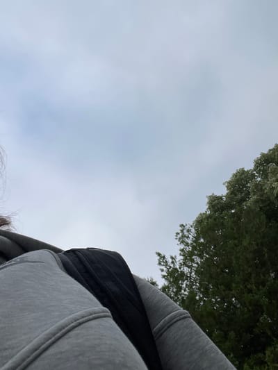 A person is wearing a grey hooded sweatshirt with a black bag visible over their shoulder. The shot is taken from a low angle, looking up towards an overcast sky. To the right of the frame, the tops of green trees are visible, suggesting an outdoor setting. The person's hair is also visible on the left side of the image. The scene appears to be in an urban or suburban environment, possibly in the city of Dobrich, Bulgaria, given the reference to that location.