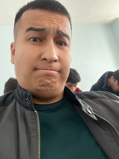 The image depicts an indoor scene, likely within a classroom or lecture hall in the city of Urgench, Uzbekistan. The primary subject is a young adult male, prominently positioned in a close-up, facing the camera. He has short, dark hair and an expression conveying mild skepticism or a deliberate pose, characterized by slightly raised eyebrows and pursed lips with a creased chin. He wears a dark grey/black zippered jacket over a dark teal/green ribbed-collar shirt. In the blurred background, two other male individuals are partially visible; one to the viewer's right wears a dark maroon garment and looks downwards, while another to the viewer's left wears a dark jacket and also looks downwards. The background consists of a plain, light blue or light teal wall, suggesting an institutional setting. The individuals in the background appear to be seated and engaged in an activity not directly involving the camera or the foreground subject.