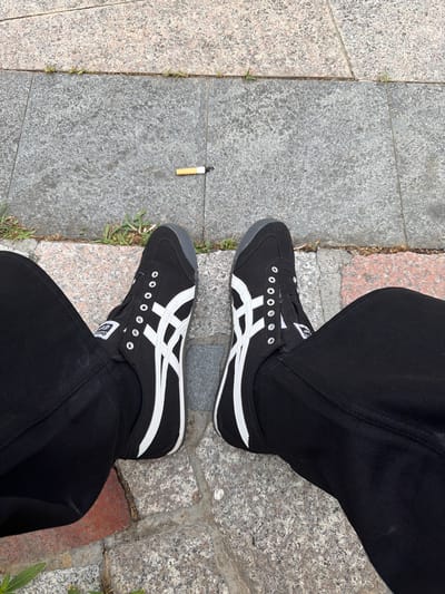The image shows a pair of black and white Onitsuka Tiger sneakers on a person's feet, seated on a stone-tiled outdoor surface. A cigarette butt is visible on the ground between the two shoes. The person is wearing black pants. The environment appears to be a public walkway, possibly in a park or street. The tiles are a mix of grey and reddish-brown granite.