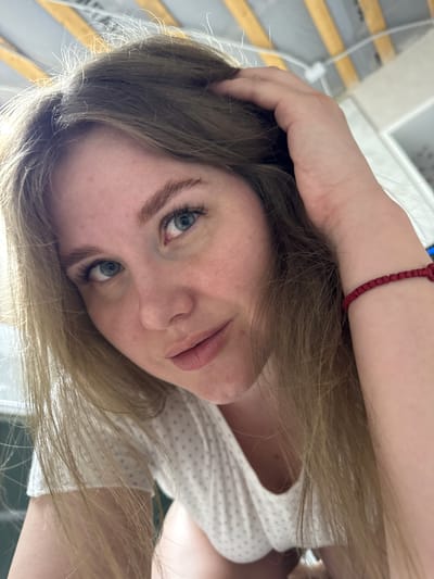 A close-up, slightly low-angle photograph depicts a female subject indoors. The subject, with long light brown hair and blue eyes, looks directly at the camera with a neutral expression. Her right hand is raised to her hair on the right side of her head. A red string bracelet is visible on her left wrist. She is wearing a white garment with faint light purple polka dots. The background above her features a ceiling structure composed of light-colored panels interspersed with parallel light brown or wooden beams. Other indistinct light-colored interior elements are visible in the periphery. The location is specified as Gornyi (Dvurechenskoe S/P), Russia.
