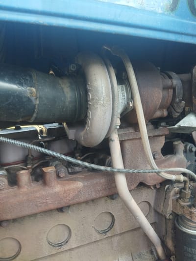 A close-up, outdoor shot depicts a section of a large, rusty engine, likely from a tractor or heavy machinery, under a blue, painted metal casing. The engine is dominated by a turbocharger assembly, with prominent silver and rust-colored metallic components. A thick, black intake pipe connects to the turbocharger's compressor housing, which has faint lettering, possibly a manufacturer's mark. Two flexible hoses, one light-colored and ribbed and another lighter, smooth hose, connect to the turbocharger. Below the turbocharger, the engine block displays a rough, textured, light brown surface with circular indentations. A rusty exhaust manifold with two protruding pipes is visible. A coiled metal wire runs horizontally across the lower portion of the image, just above the engine block. The background is dark and out of focus, with the blue metal casing forming the top edge. No people are present, and the scene appears to be in a state of disrepair or storage. The location is identified as Petroplavlovka, Kyrgyzstan.
