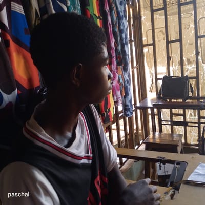 In Nsugpe, Nigeria, a young dark-skinned man sits at a sewing machine. He is wearing a white t-shirt with a black and red striped overlay. To his left, fabrics hang on a rack. To his right, a window with metal bars is visible; a woman's face appears to be visible behind the bars. A black bag sits on a table near the window.