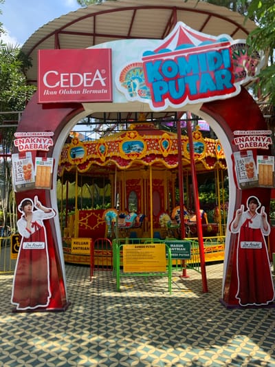 The image shows a carousel in the middle of the scene, with a red, yellow, and blue color scheme. The carousel is surrounded by a green fence with yellow and white warning signs, and there is a sign with the Indonesian text "KOMIDI PUTAR". There is a red archway on each side of the carousel, with a red and white sign that says "CEDEA Ikan Olahan Bermutu" and "CEDEA KOREAN FOOD ENAKNYA DAEBAK" on one side, and "CEDEA KOREAN FOOD ENAKNYA DAEBAK" on the other side. The signs on both sides also feature images of women dressed in traditional Korean clothing. There is green foliage and a blue sky in the background. The carousel appears to be at an amusement park located in Batu, Indonesia, at coordinates -7.884 latitude and 112.525 longitude. The carousel is stationary and there are no people interacting with it.  The ground is tiled with a patterned design in shades of beige, yellow, and green. The image is taken from an angle slightly above the carousel. The lighting is bright and the sun is casting shadows on the ground.  There is no one around the carousel or in the immediate surroundings.