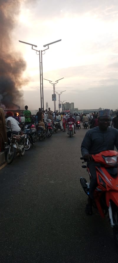 A crowd of people gathers on a road alongside multiple motorbikes. Dark smoke billows in the background near some buildings, indicating a possible fire. A man on a red motorbike faces the camera. Overhead, street lights line the road above the crowd. The sky is overcast, suggesting the picture was taken either early morning or late afternoon.