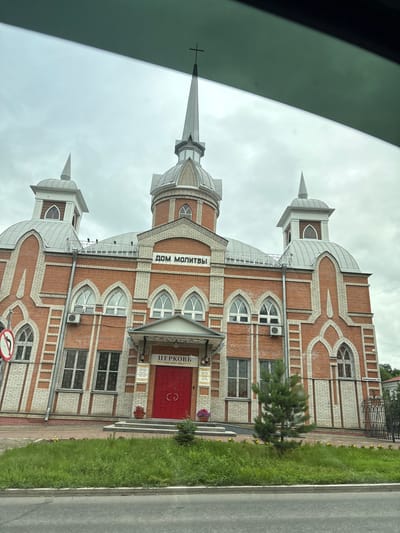The image shows a brick building with a large domed roof and a spire, likely a church or religious building. The building is adorned with white decorative brickwork. There are several arched windows and a prominent red double door entrance. Above the entrance, there is a sign with Russian text that appears to read "ЦЕРКОВЬ" (Church). Another sign above this reads "ДОМ МОЛИТВЫ" (House of Prayer). The building is situated in a grassy area with some small trees. A street is visible in the foreground. The sky is overcast. The building is located at ul. Leningradskaya, 22a in Khabarovsk, Russia, and is identified as the Church of the Resurrection of Christ.