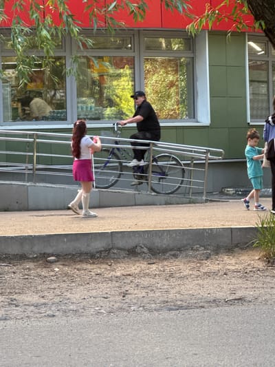 A group of people are seen on a paved area in front of a building with a red awning and green walls. A man wearing a black shirt and cap is riding a bicycle on a ramp. To his left, a young woman with long brown hair, wearing a white shirt and pink skirt, is holding a pink object. To the right of the man on the bicycle, a young boy in a turquoise shirt and shorts is walking, carrying a white bag. In the background, a window reflects trees and sunlight. The scene appears to be outdoors in a developed area.