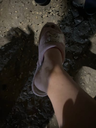 A close-up, overhead shot shows a person's right foot wearing a light pink slipper with a textured, ribbed design and a white applique on the strap. The foot is positioned on a rough, uneven surface composed of dark soil, gravel, and small stones. Shadows are cast across the ground, suggesting artificial lighting and an outdoor setting, possibly at night. The person's leg, from the ankle upwards, is visible in the frame, appearing to be light-skinned. The background is a mixture of dark, granular material and lighter-colored stones. No visible text is present in the image. The scene is set in Verin Artashat, Armenia.