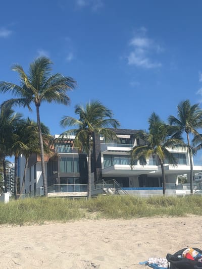 A modern multi-story beach house with large windows and balconies is situated on a sandy beach in Fort Lauderdale, Florida. Several palm trees are in the foreground, with one prominently in the center, obscuring parts of the house. The sky is clear blue with a few scattered clouds. The lower level of the house appears to have an infinity pool with blue tile. In the bottom right corner of the image, a black bag and some clothing items are visible on the sand.