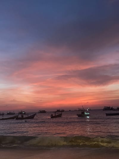 The image displays a coastal sunset scene in Ko Tao, Thailand. The sky presents a color gradient, transitioning from deep indigo and purple at the top to vibrant reds, oranges, and pinks across the mid and lower sections, with illuminated stratiform clouds. The water, dark and reflecting the sky's hues, contains approximately twenty vessels, predominantly small fishing or longtail boats, distributed across the visible expanse. Most boats are dark silhouettes, while several exhibit active lights, casting distinct reflections on the water's surface. One boat, positioned mid-ground to the right of center, features a prominent green light. The immediate foreground shows a sandy shoreline with a visible breaking wave, indicating an active tide. No human figures or specific interactions are discernible.