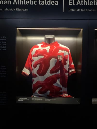 This media file captures a display of a red and white patterned football jersey, likely belonging to Athletic Club. The jersey is presented in a display case, illuminated from within, highlighting its intricate design. The backdrop suggests a museum or exhibition setting, possibly dedicated to the history of Athletic Club.

Visible text on the dark blue wall to the left of the display reads "oen Athletic taldea", which translates from Basque to "Athletic Team". Further down, "e nahasiak Klubean" is visible, meaning "mixed in the Club". Text on the right side of the display, though partially obscured and rotated, includes "Debut de las Leonas", indicating a debut for "the Lionesses," a common nickname for women's sports teams. Other fragmented text in Spanish refers to a "Camiseta vollenca" and "D. Ursay mangolariaren" and "platu nuen", possibly referencing the jersey's designer or a significant event associated with it.

The overall impression is one of historical preservation and celebration of athletic heritage. The lighting and presentation create a sense of reverence for the displayed artifact. The context provided by the text suggests a focus on the evolution and significant moments of the Athletic Club, potentially including its women's team. The setting appears to be indoors, with controlled lighting, making it difficult to determine the time of day or weather.